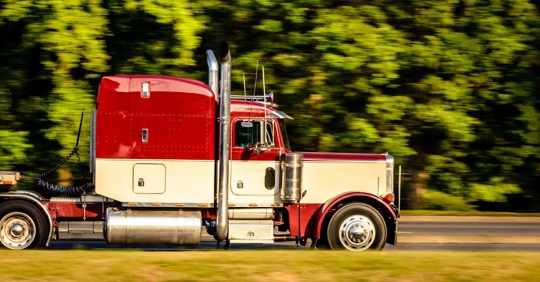 red semi-truck driving past a line of trees