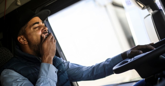young man driving a large truck, covering his mouth with his hand while yawning