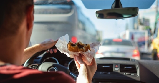 man sitting in the driver's seat of a car in a traffic jam holding a half-eaten burger in his right hand