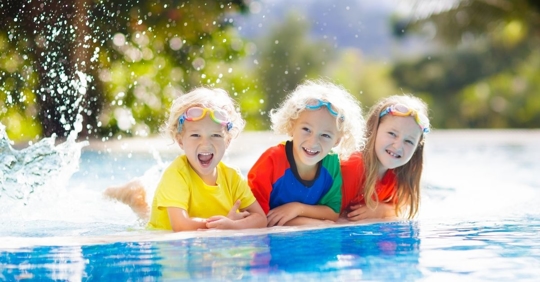 three blonde children laying on the edge of a swimming pool and smiling